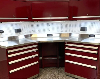 Corner industrial workstation with stainless steel work surface, red drawer cabinets, overhead storage and pegboard for tool organization.