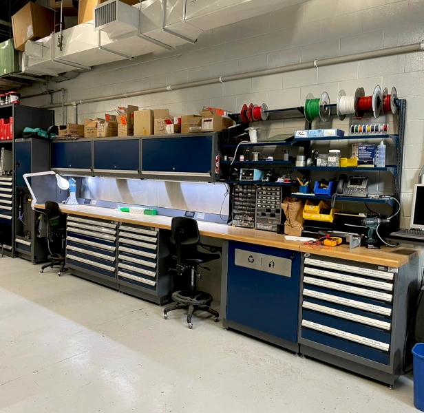 Industrial workstation with long wooden work surface, integrated tool cabinets, overhead storage and ergonomic setup for electronics and assembly tasks.